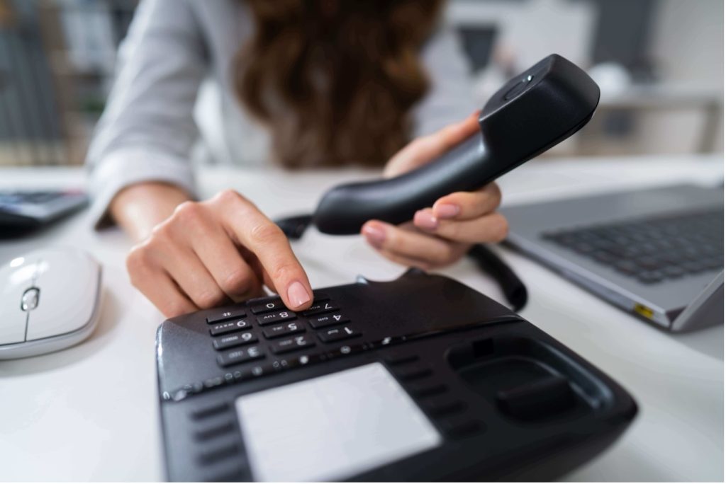 On aperçoit une femme assise à son bureau qui compose un numéro sur son téléphone fixe. 